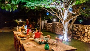 An outdoor dining area with a wooden table set for a meal, surrounded by trees and stone walls. - Aruba Boutique Apartments Adults Only (Noord)