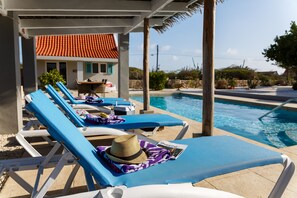 A poolside area with lounge chairs, a pool, and a building with a red-tiled roof in the background. - Aruba Boutique Apartments Adults Only (Noord)