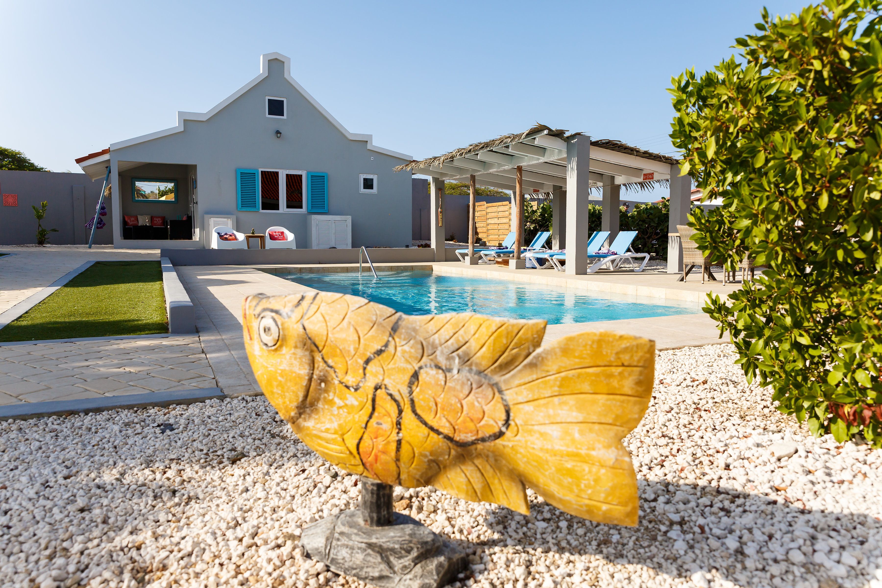 Piscina com espreguiçadeiras, edifício com persianas azuis e uma escultura decorativa de peixe.