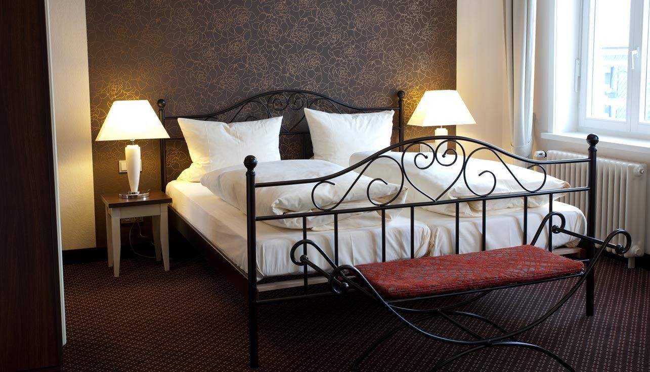 A neatly made bed with white linens and two bedside lamps, a red cushioned chair, and a patterned wall.