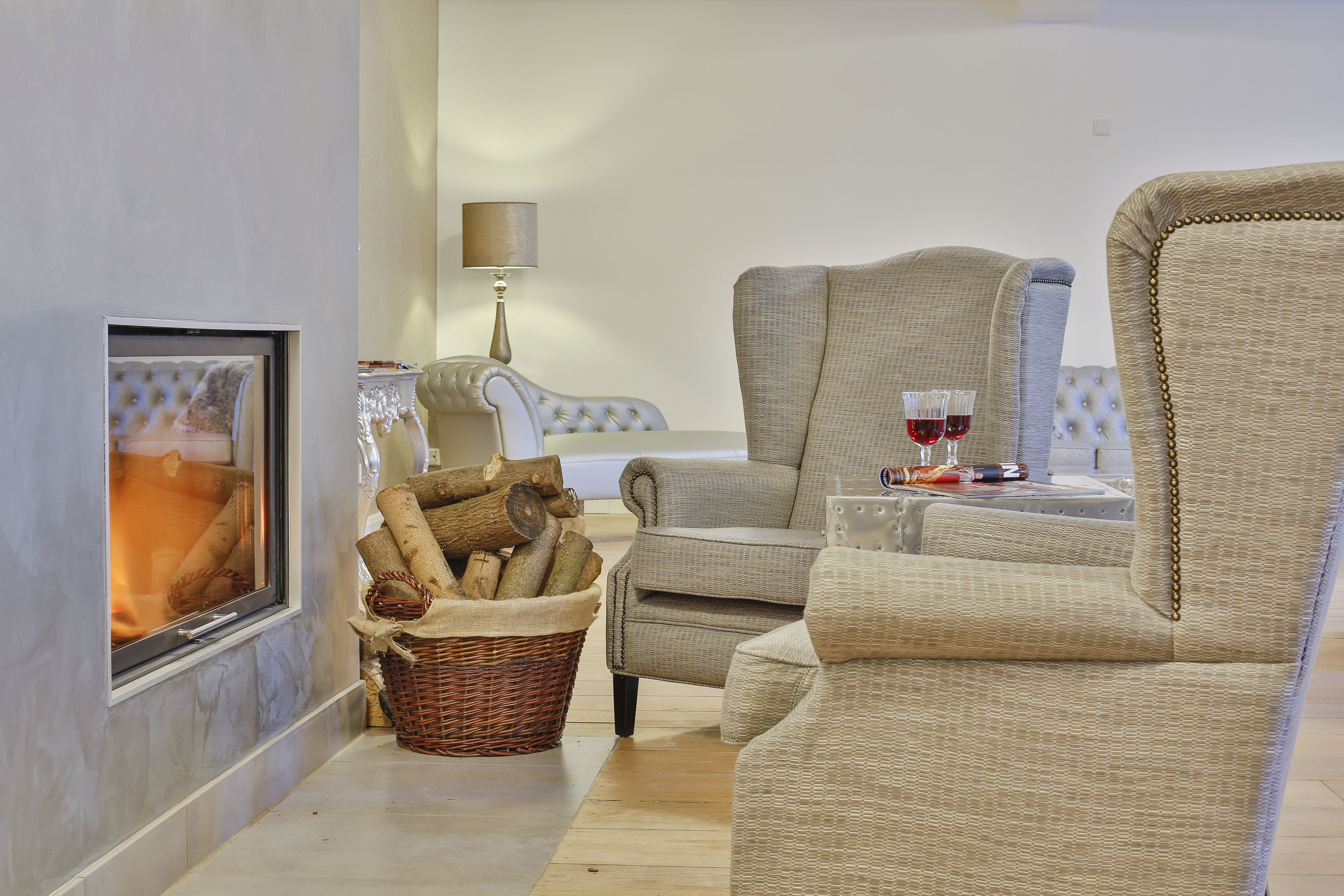 A cozy living room with a fireplace, a wicker basket with logs, a lamp, and two chairs with glasses of red wine.