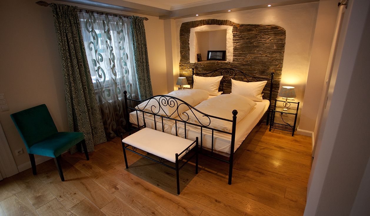A bedroom with a double bed, a chair, a bedside table with a lamp, and a stone wall.