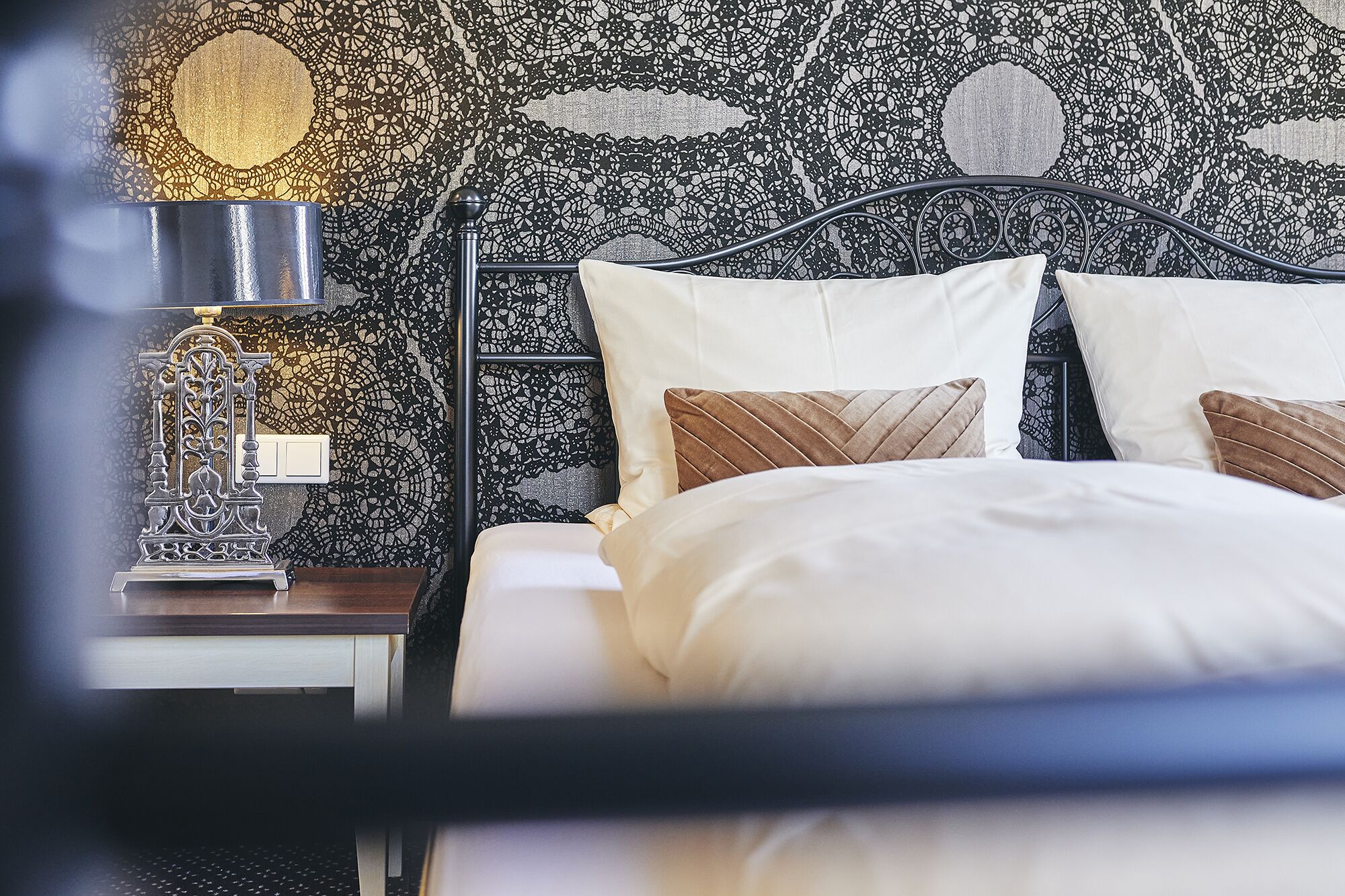 A neatly made bed with white and brown pillows, a black metal headboard, and a nightstand with a lamp.