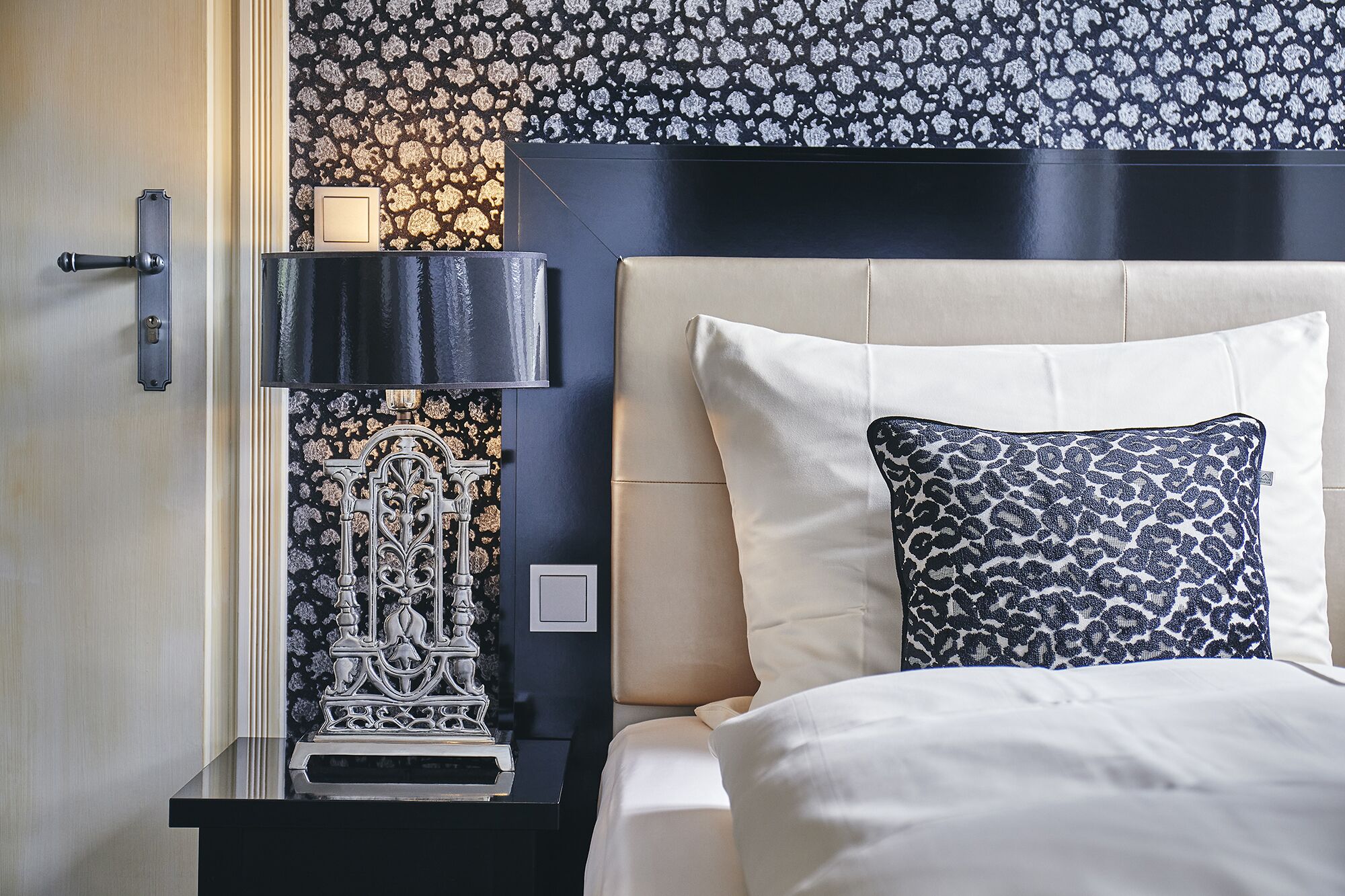 A neatly made bed with a white and blue patterned pillow, a black nightstand with a lamp, and a decorative wall piece.