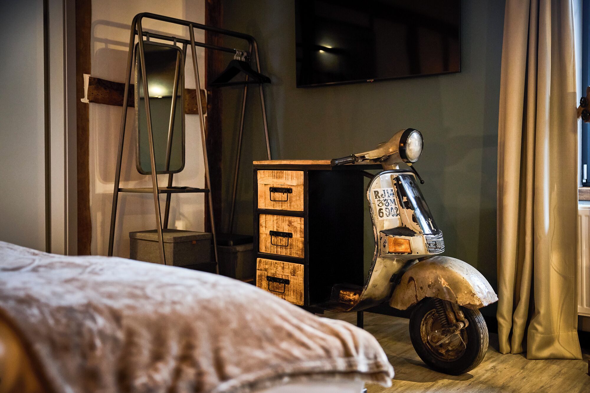 A room with a vintage scooter, a wooden chest of drawers, a metal chair, and a bed.