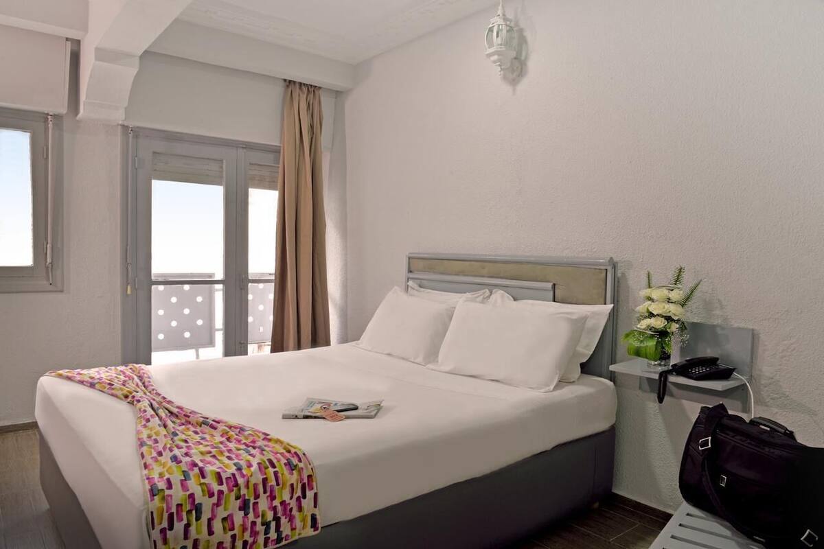 Une chambre d’hôtel avec un lit, une table de chevet, une lampe, une fenêtre avec des rideaux et une vue sur le ciel.