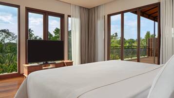 A large bed with a TV on a wooden stand, a balcony with a view of greenery, and a large window with curtains.
