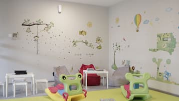 Una sala de juegos infantil con un tapete acolchado, dos mecedoras con forma de rana, una mesita blanca y un sillón de puf rojo.