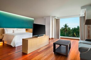 Habitación de hotel moderna con una cama grande, un televisor sobre un mueble de madera, una mesita pequeña y vistas a palmeras a través de amplios ventanales.