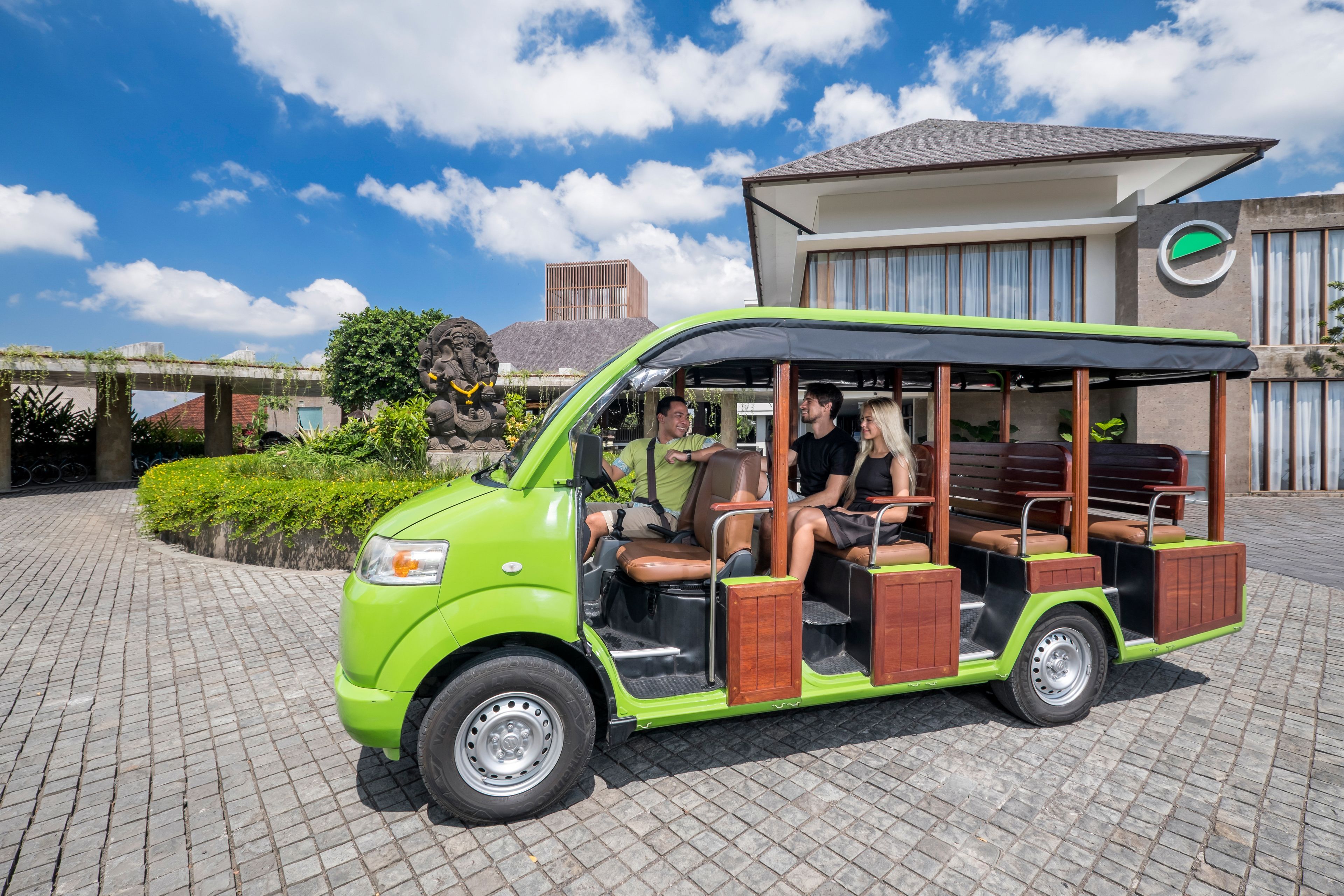 Een groene en zwarte trolley met passagiers, geparkeerd voor een gebouw met een modern ontwerp en een stenen beeldhouwsel.