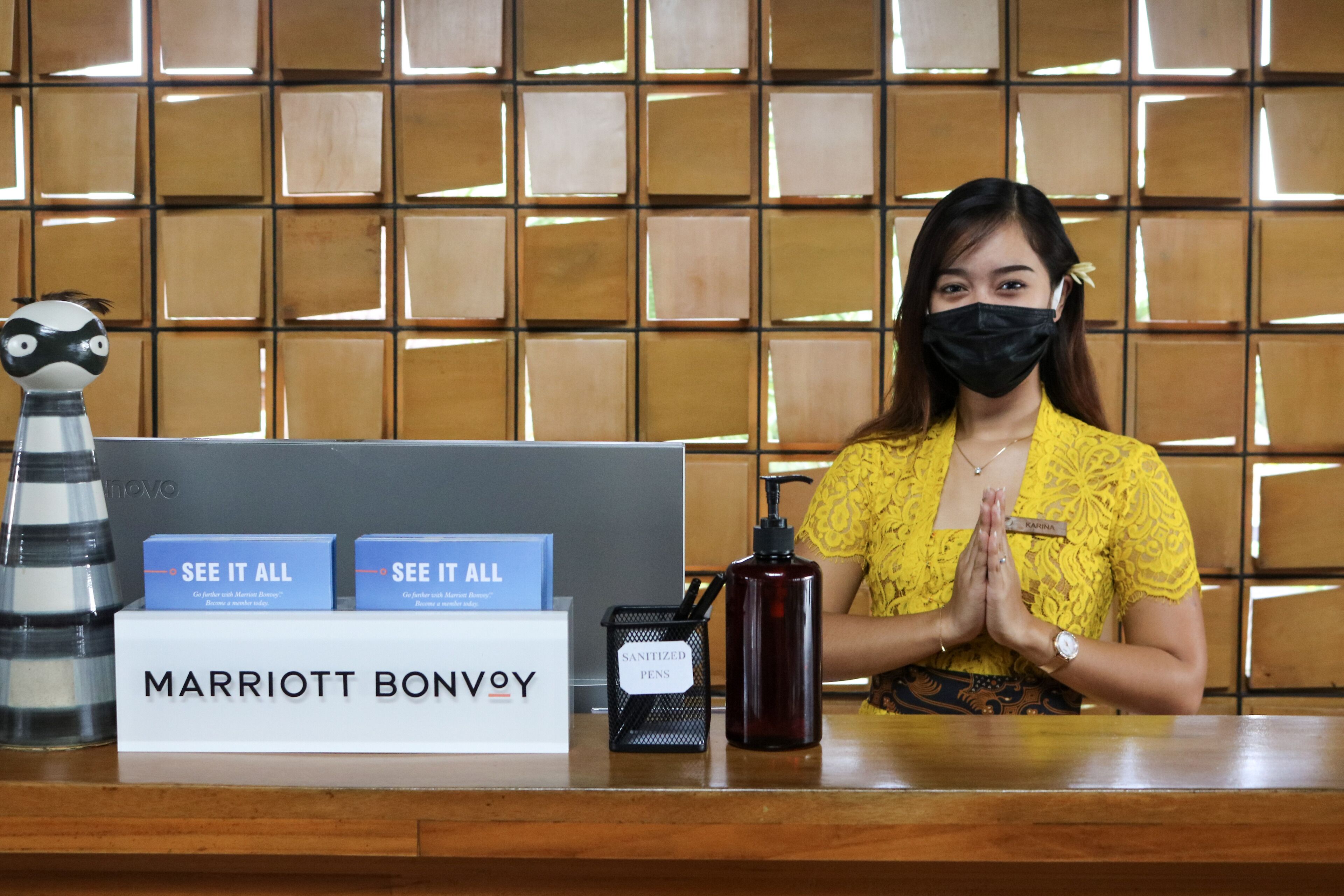 Un mostrador de recepción con un cartel que dice "MARRIOTT BONVOY" y una identificación con el nombre "KARINA". Hay dos letreros que dicen "SEE IT ALL", una botella de agua y un dispensador de desinfectante.