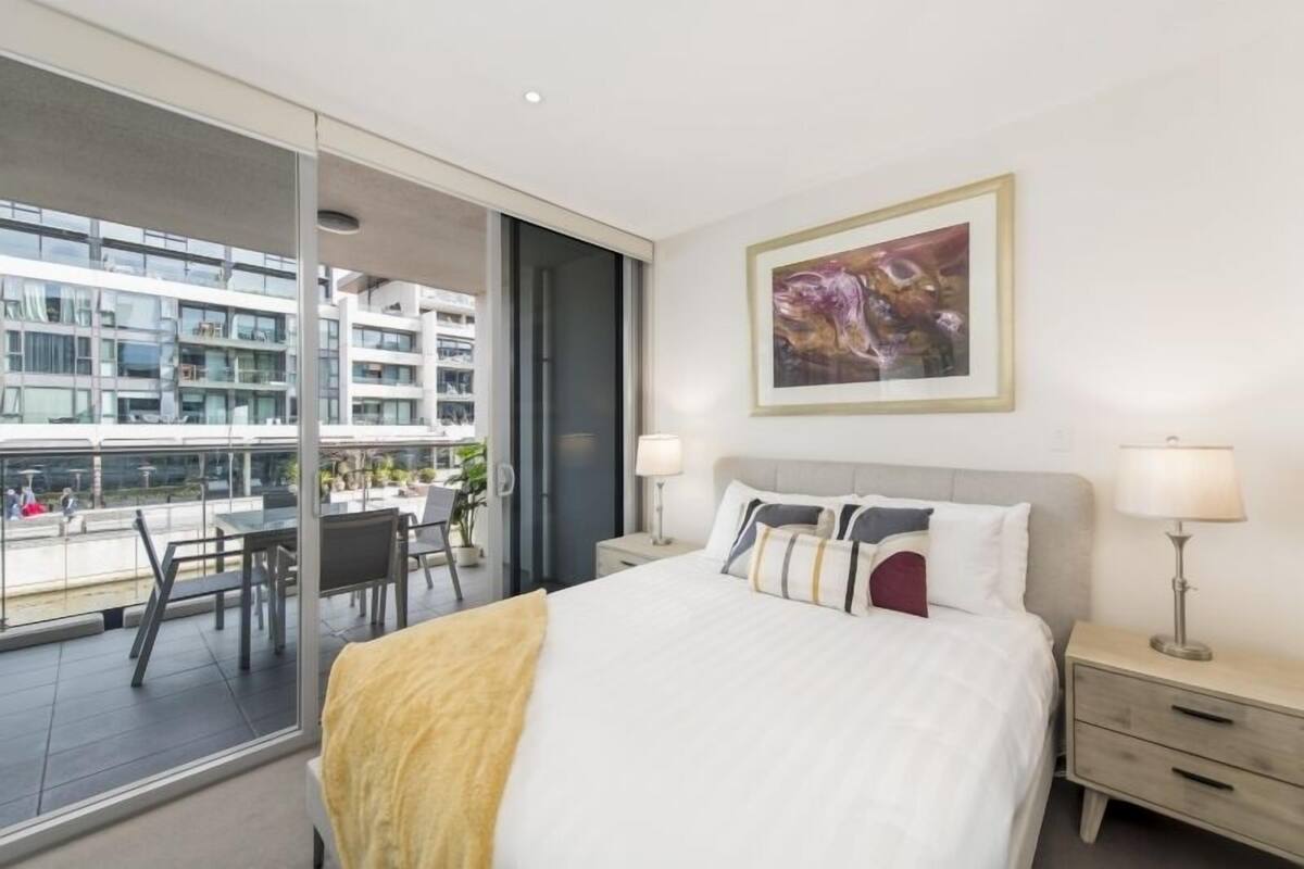 A hotel room with a bed, two bedside tables, a large window, and a view of a modern building.