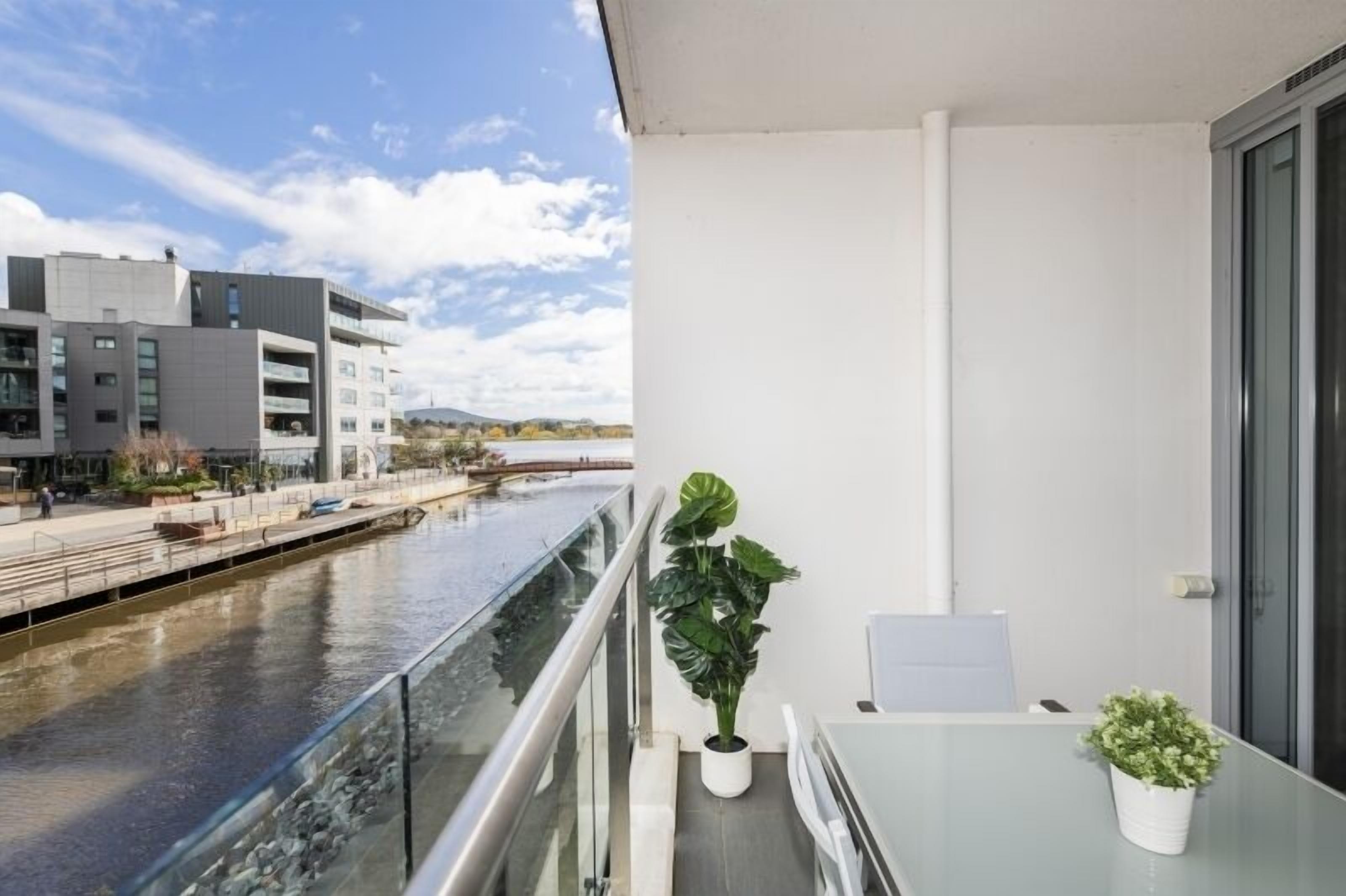 A balcony with a glass railing overlooking a river, a potted plant, and a small table.
