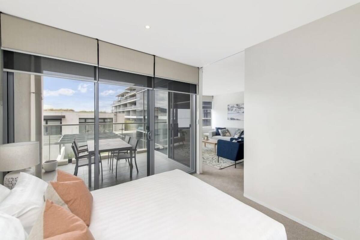 A modern hotel room with a large bed, a balcony with a dining area, and a view of the cityscape.