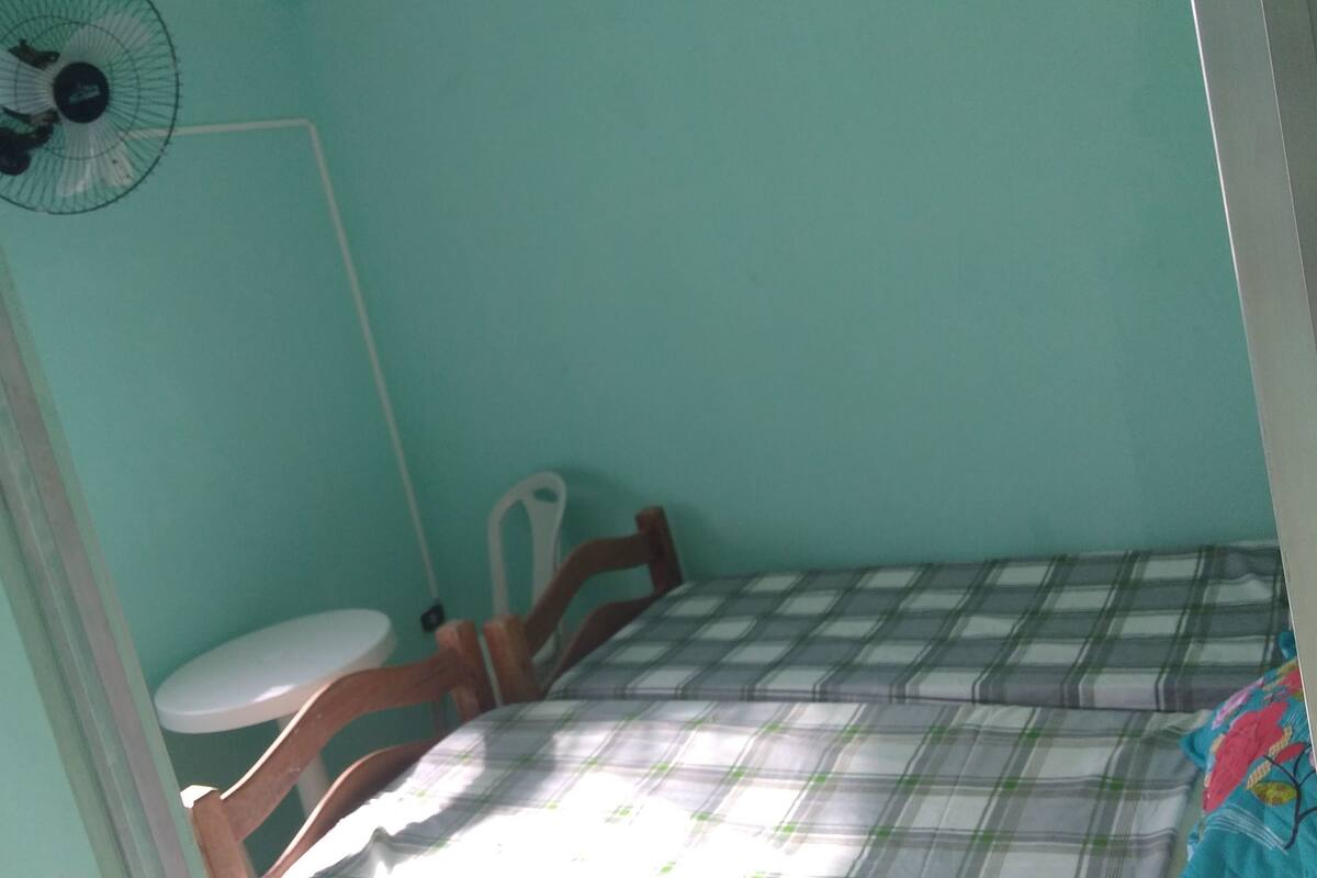 A small, single-bed room with a wooden bed frame, a white toilet, and a wall-mounted fan.