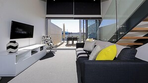 A modern living room with a flat-screen TV, a sofa with yellow pillows, a patterned armchair, and a glass wall offering a city view. - Accommodate Canberra - Mode (Braddon)