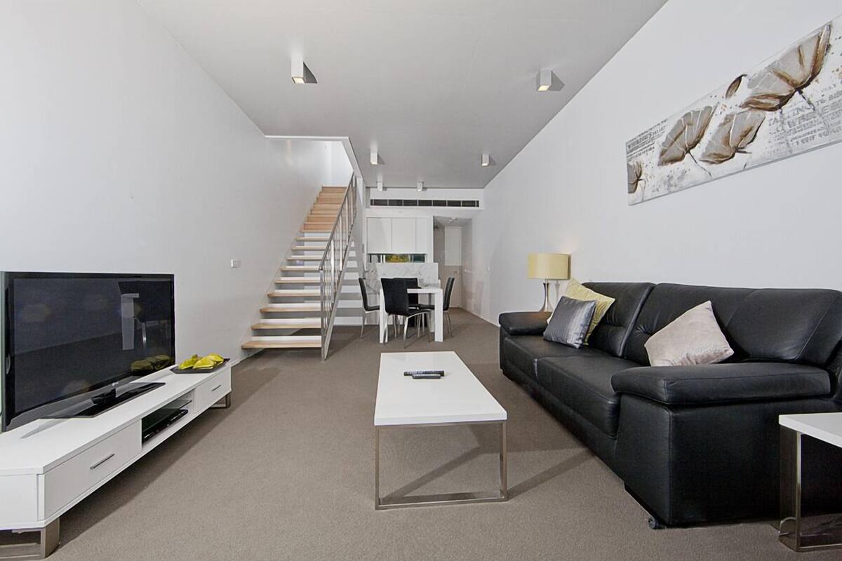A modern living room with a flat-screen TV, a black leather sofa, a white coffee table, and a staircase leading to an upper level.
