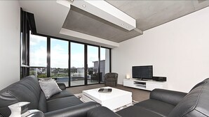A modern living room with a black leather sofa, a white coffee table, and a flat-screen TV on a white console. Large windows offer a city view. - Accommodate Canberra - The ApARTments (Canberra)