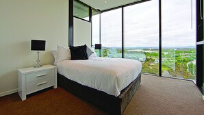 A bedroom with a large bed, a nightstand, a lamp, and a view of a cityscape through floor-to-ceiling windows. - Accommodate Canberra - The ApARTments (Canberra)