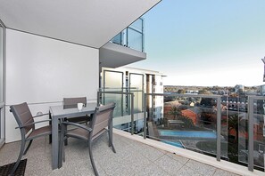 A modern balcony with a glass railing, a dining table with chairs, and a view of a swimming pool and buildings in the background. - Accommodate Canberra - The Avenue (Turner)