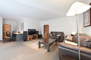 A modern living room with a sofa, coffee table, TV, and a desk with a computer. There are bookshelves and a lamp. - Accommodate Canberra - The Avenue (Turner)
