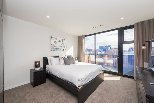 A modern bedroom with a large bed, a nightstand, a desk, and a view of the city through a large window.