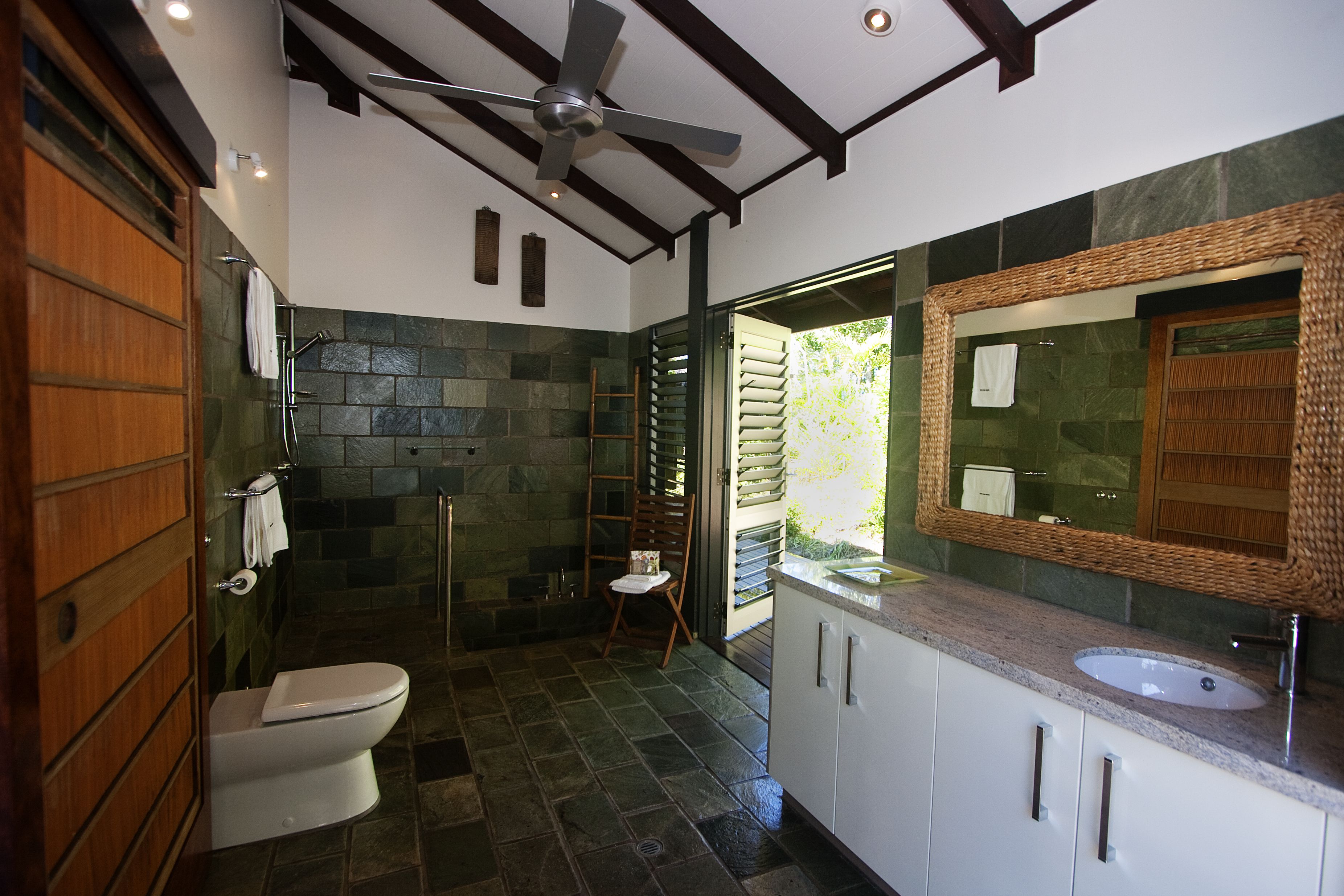 A modern bathroom with a stone floor, a white toilet, a large mirror, and a ceiling fan.