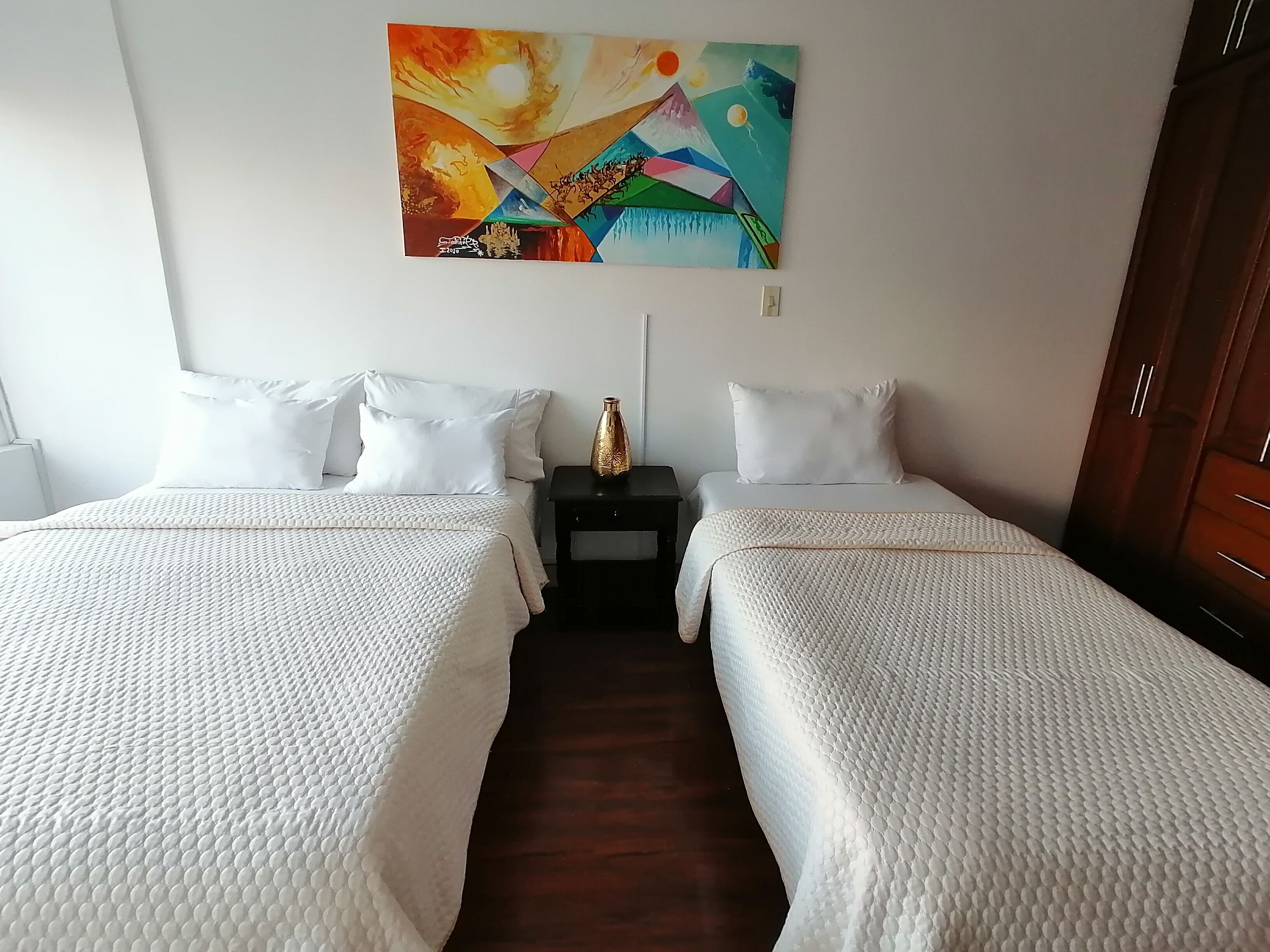Habitación de hotel con dos camas, ropa de cama blanca, una mesita de noche con un florero y un cuadro abstracto colorido en la pared.