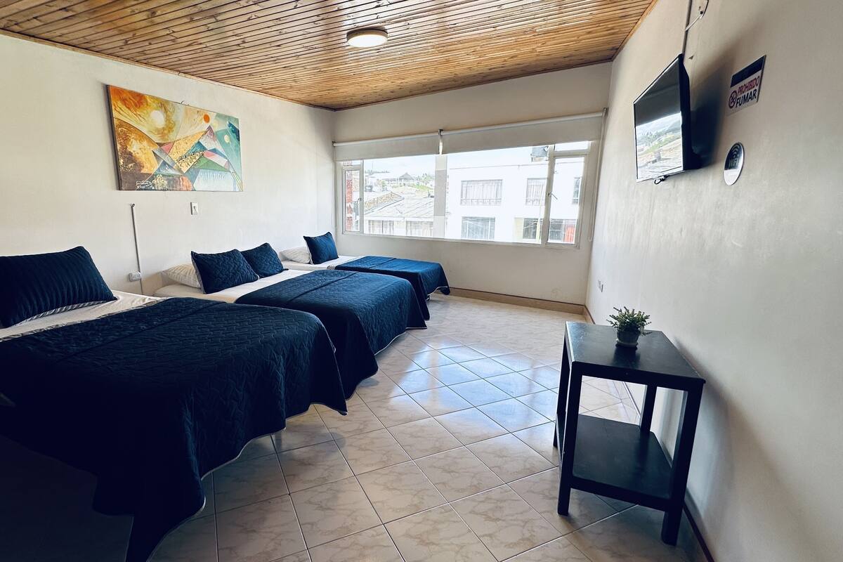A room with two beds, a wooden ceiling, a painting, a TV, and a window with a view of buildings.