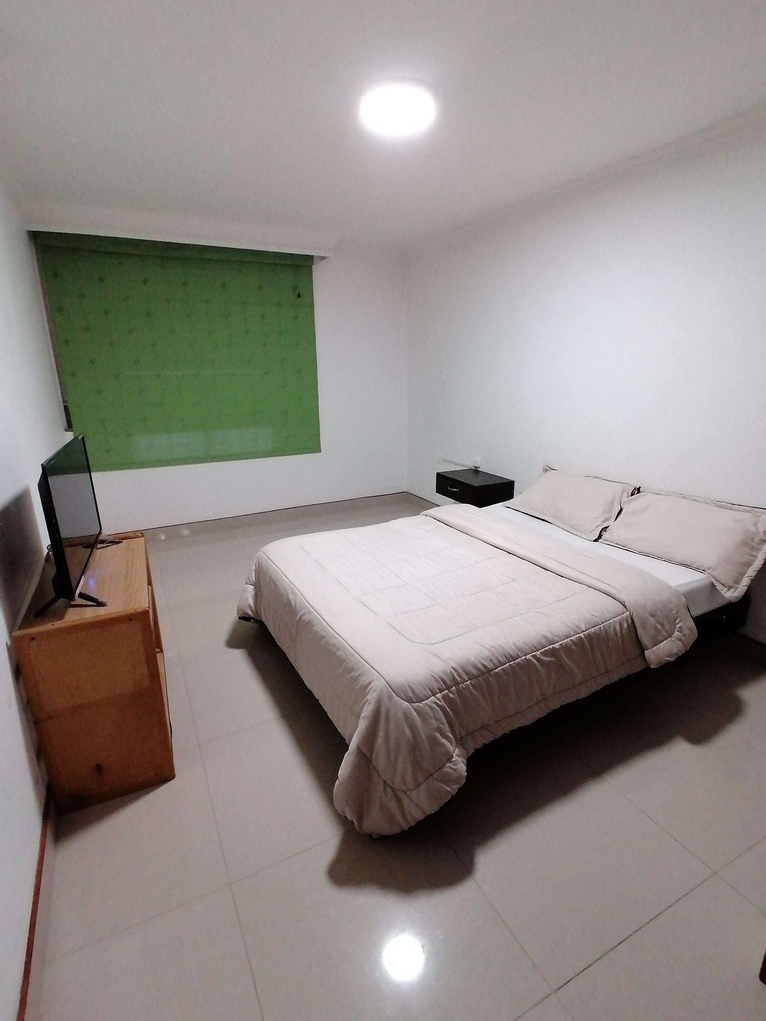 A bedroom with a bed, a nightstand, a TV on a stand, and a green window blind.