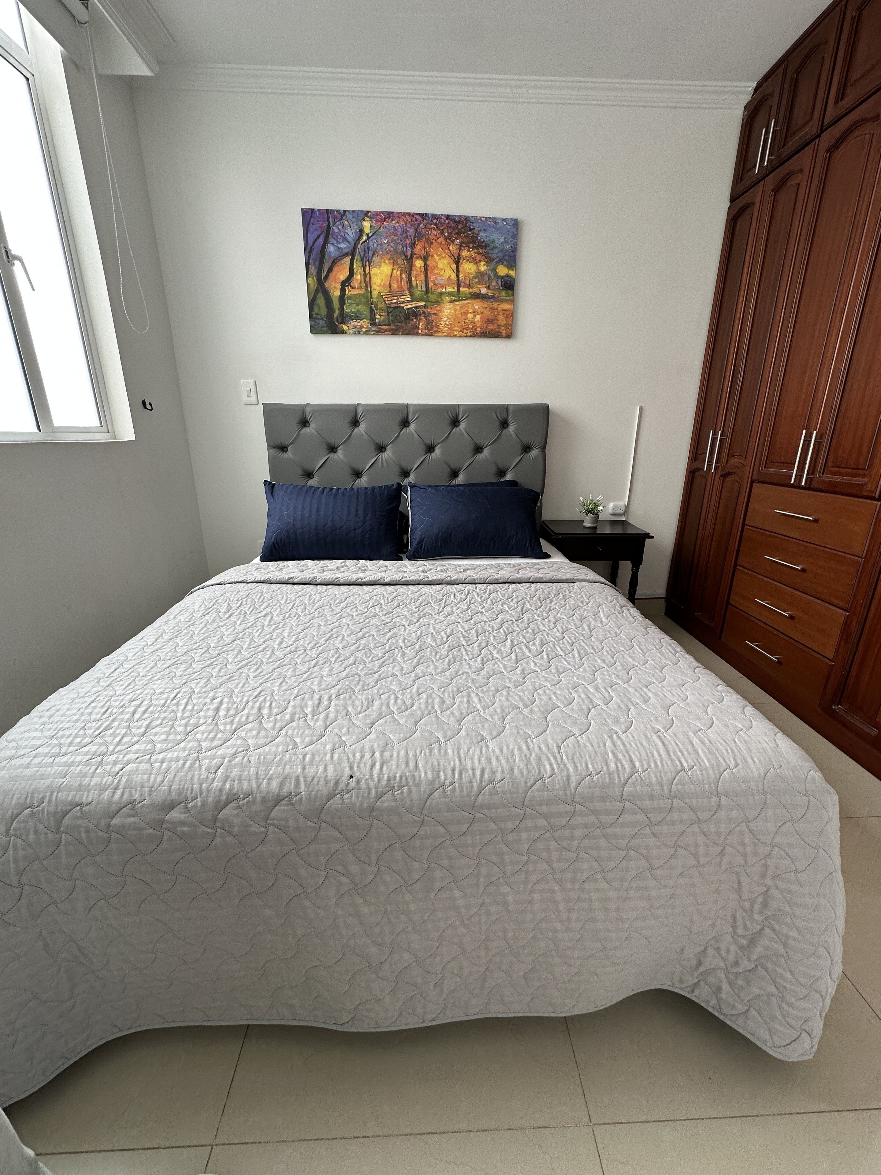 Un dormitorio con una cama grande, un cabecero tapizado gris, un cuadro en la pared y un armario de madera.
