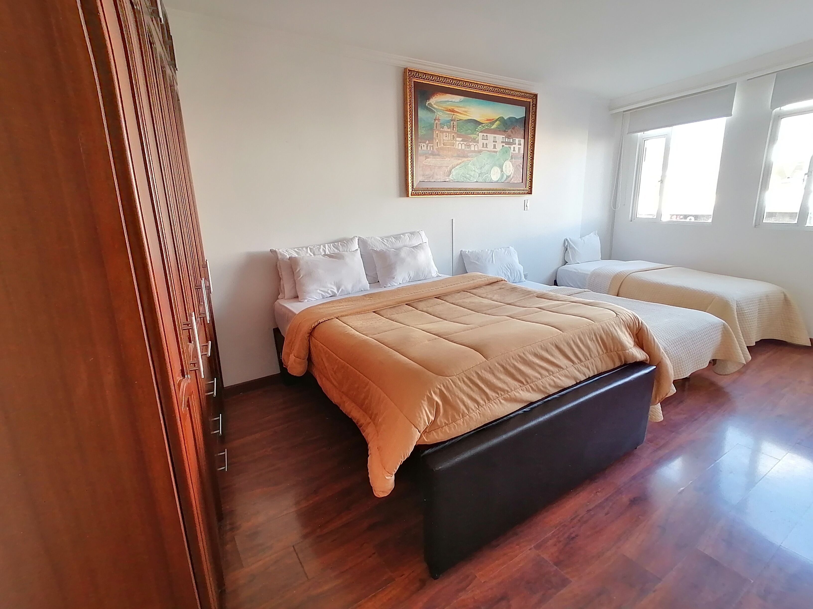 A bedroom with a large bed, two smaller beds, a wooden floor, a painting on the wall, and a window with natural light.
