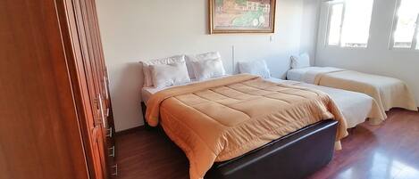 A bedroom with a large bed, two smaller beds, a wooden floor, a painting on the wall, and a window with natural light.