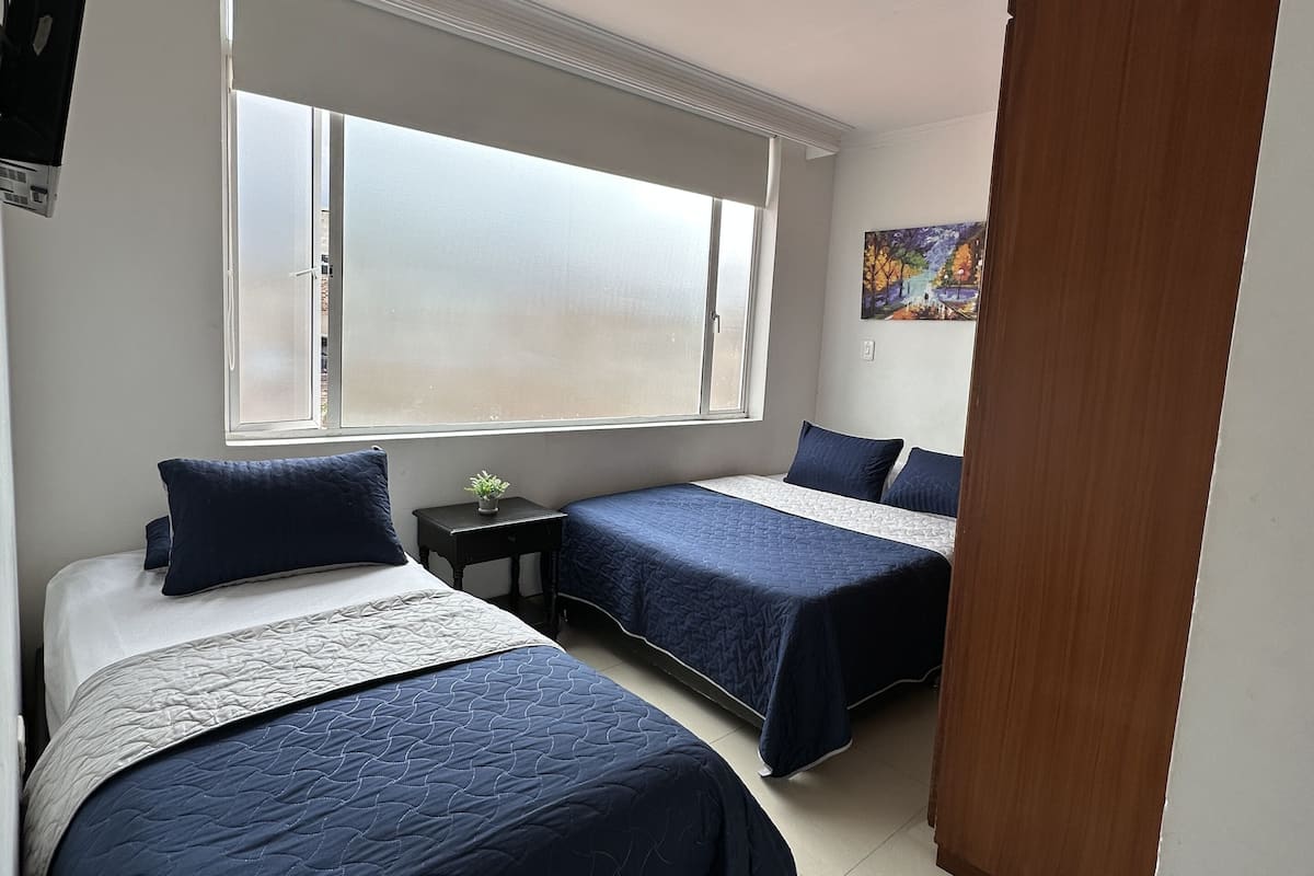 A bedroom with two beds, a large window, a painting on the wall, and a wooden door.
