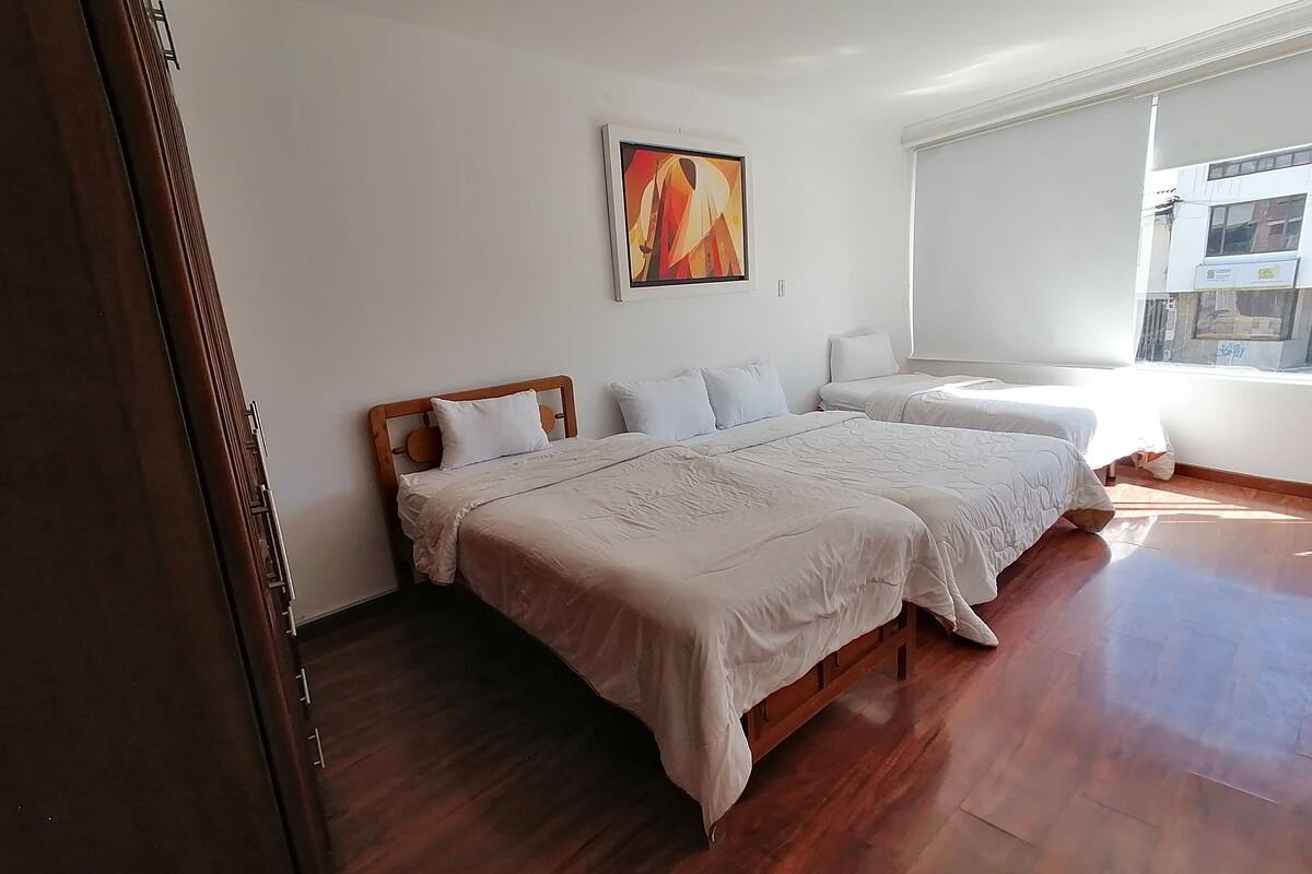 A hotel room with two beds, a wooden floor, a painting on the wall, and a window with a view of buildings.