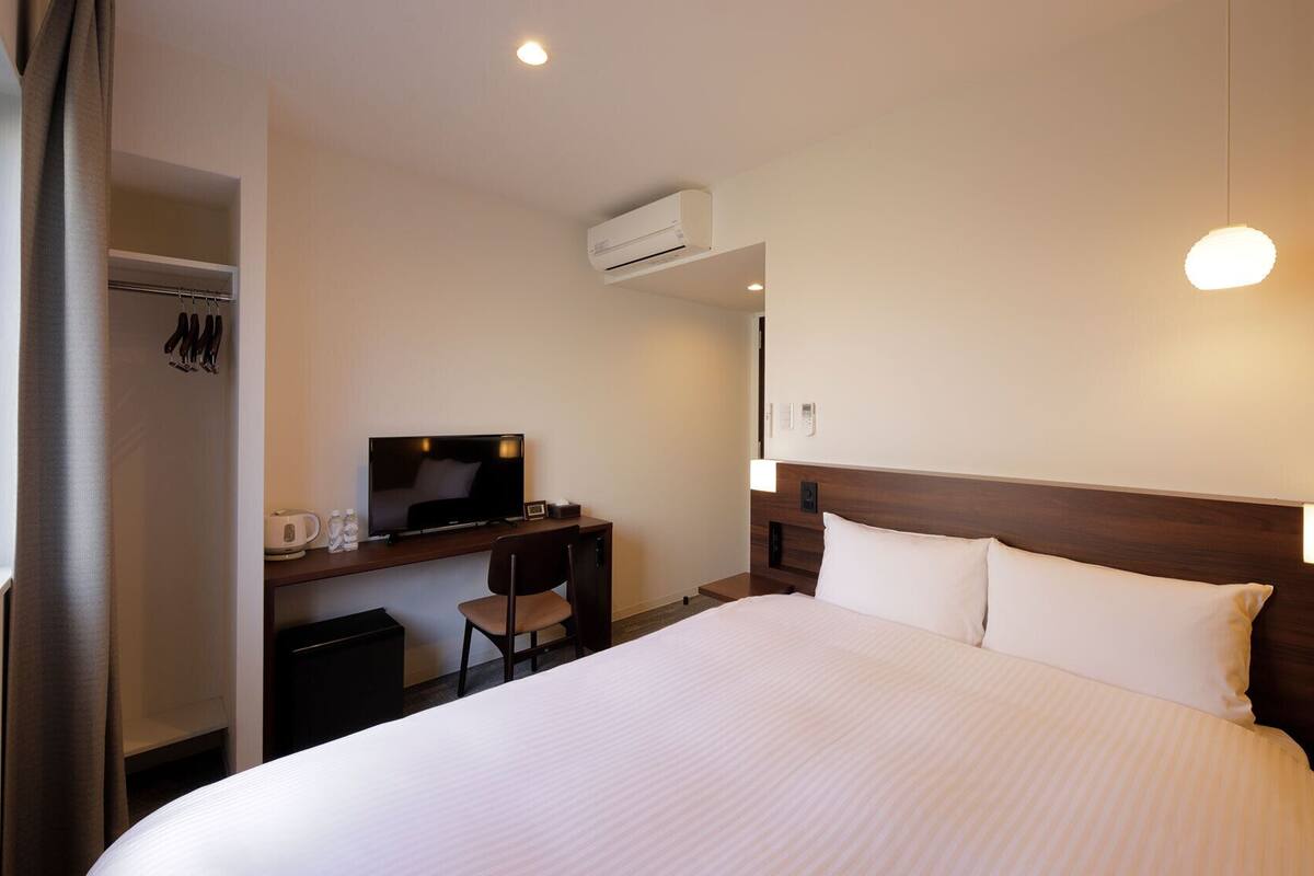 A hotel room with a bed, a desk with a TV, a coat rack, and an air conditioning unit.