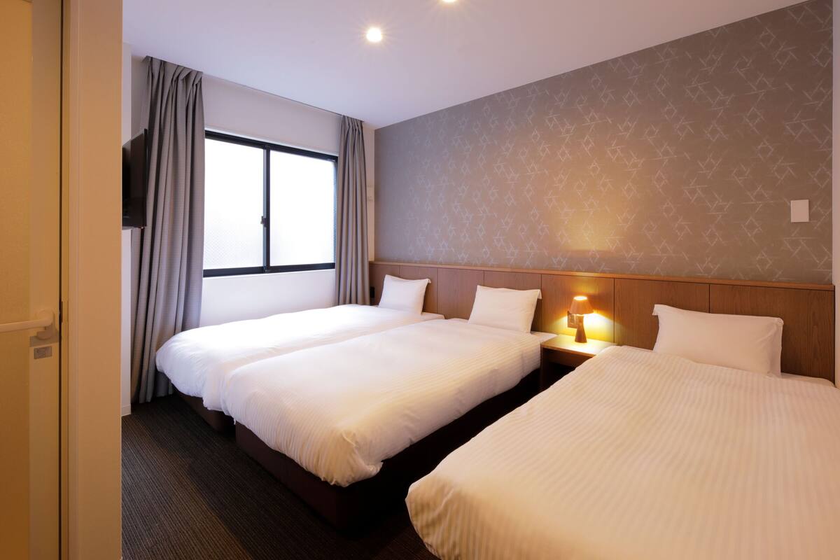 A hotel room with two beds, a TV mounted on the wall, a window with curtains, and a patterned wall.