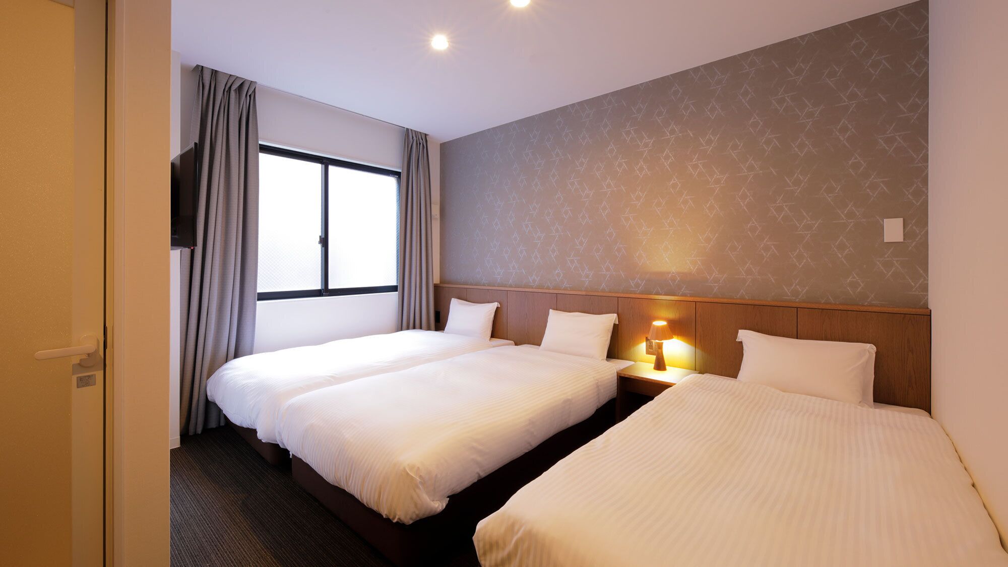 A hotel room with two beds, a window with curtains, a patterned wall, and a nightstand with a lamp.