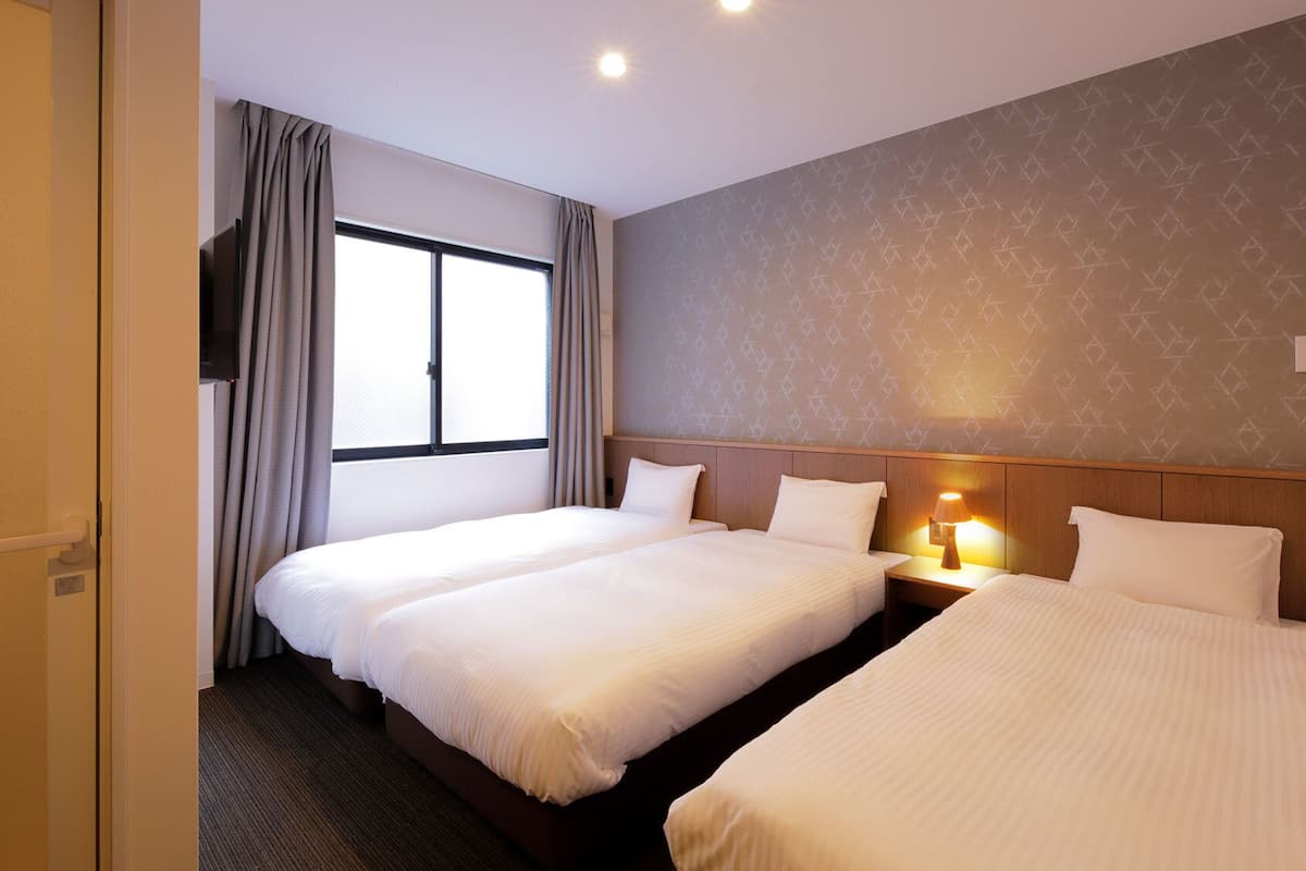 Habitación de hotel con dos camas, una ventana con cortinas, un papel tapiz estampado y una mesita de noche con lámpara.