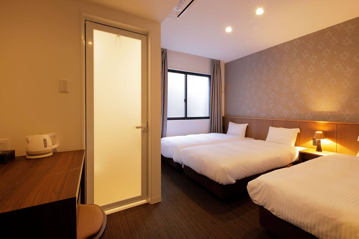 A hotel room with two beds, a desk with a kettle, a window with curtains, and a patterned wall.