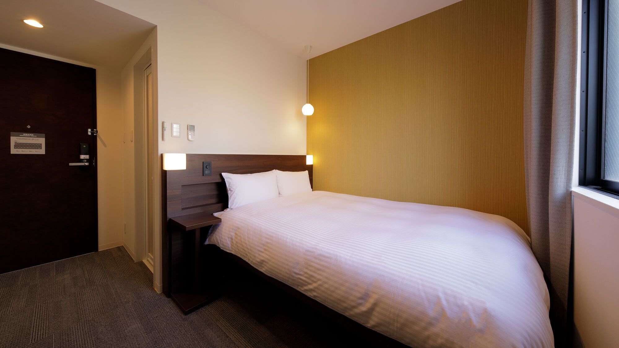 A hotel room with a bed, bedside table, lamp, and a door with a sign.