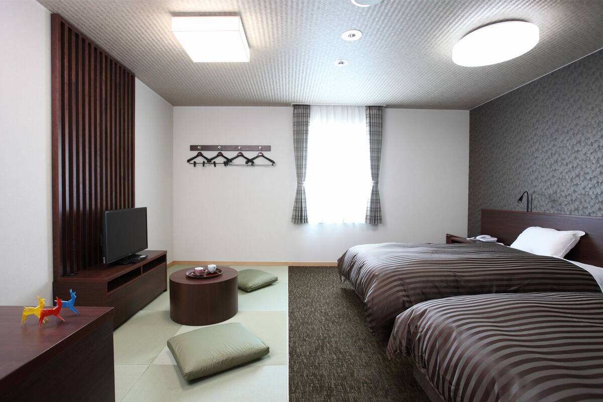 Quarto de hotel moderno com televisão de tela plana, duas camas, um pequeno espaço de estar e uma janela com cortinas.