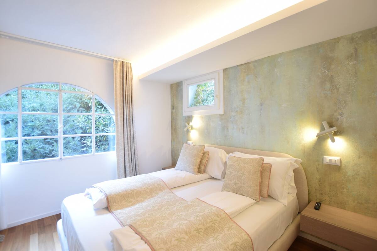 A bedroom with a large bed, a window with curtains, and a view of greenery outside.