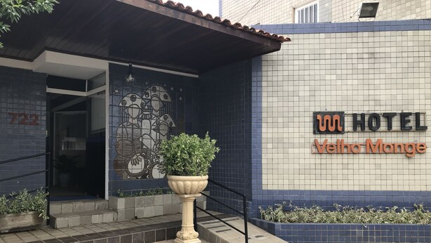 Entrada de hotel com fachada azulejada, um vaso grande com planta e uma placa com a inscrição '100 HOTEL Velho Monte'.