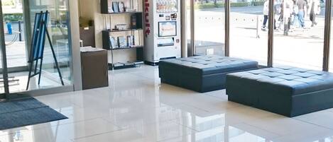 Un vestíbulo moderno con entrada de vidrio, zona de estar, máquina expendedora y un piso limpio y reflectante.