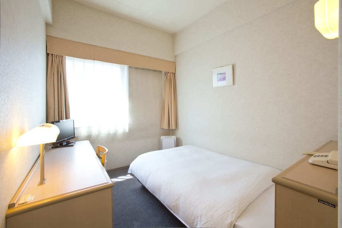 Camera d'albergo con un letto, una scrivania con lampada, una sedia, un comodino con telefono e una finestra con tende.