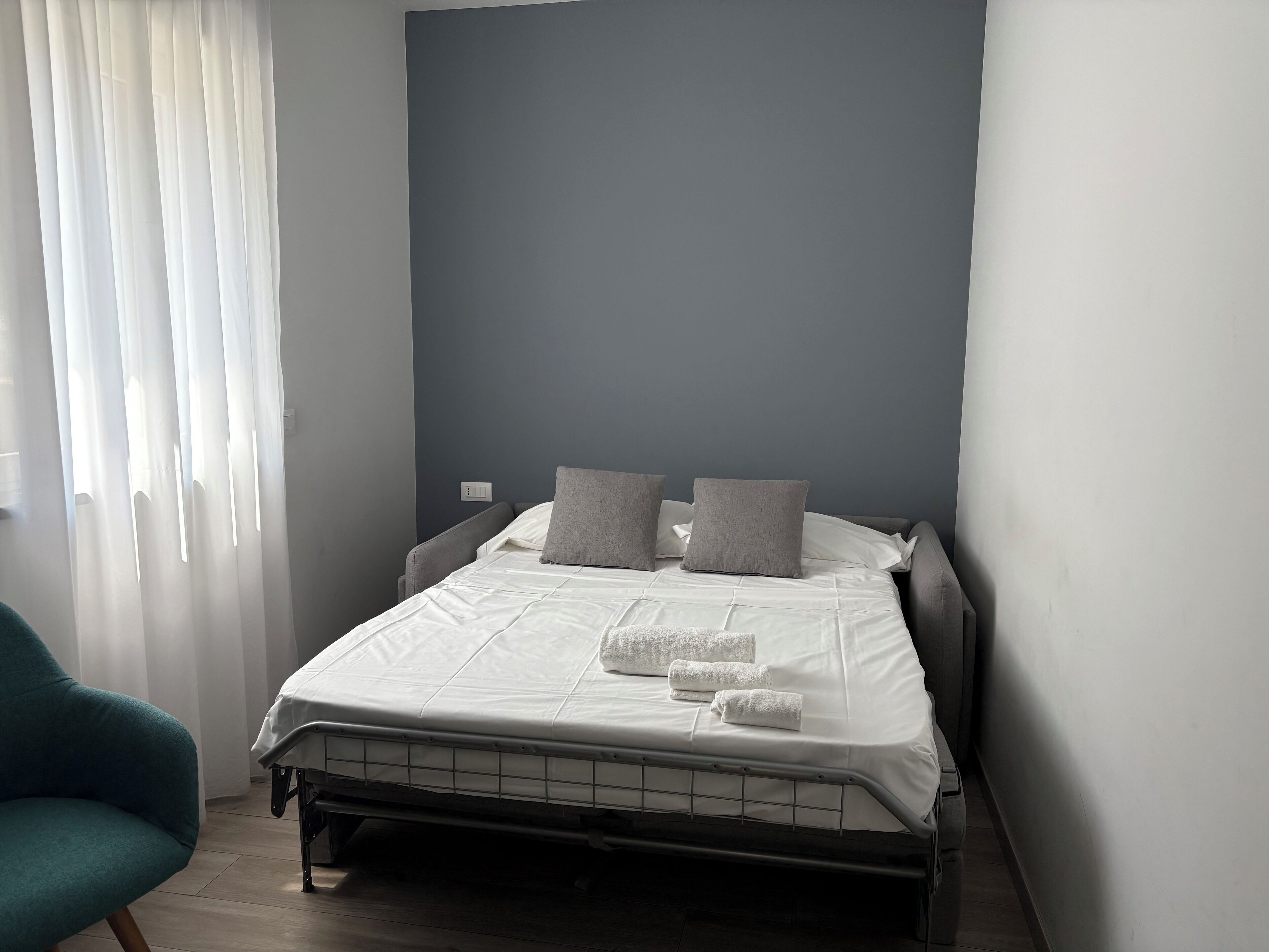 Ein ordentlich bezogenes Bett mit Kissen und Handtüchern, ein türkisfarbener Sessel und ein Fenster mit leichtem Vorhang.