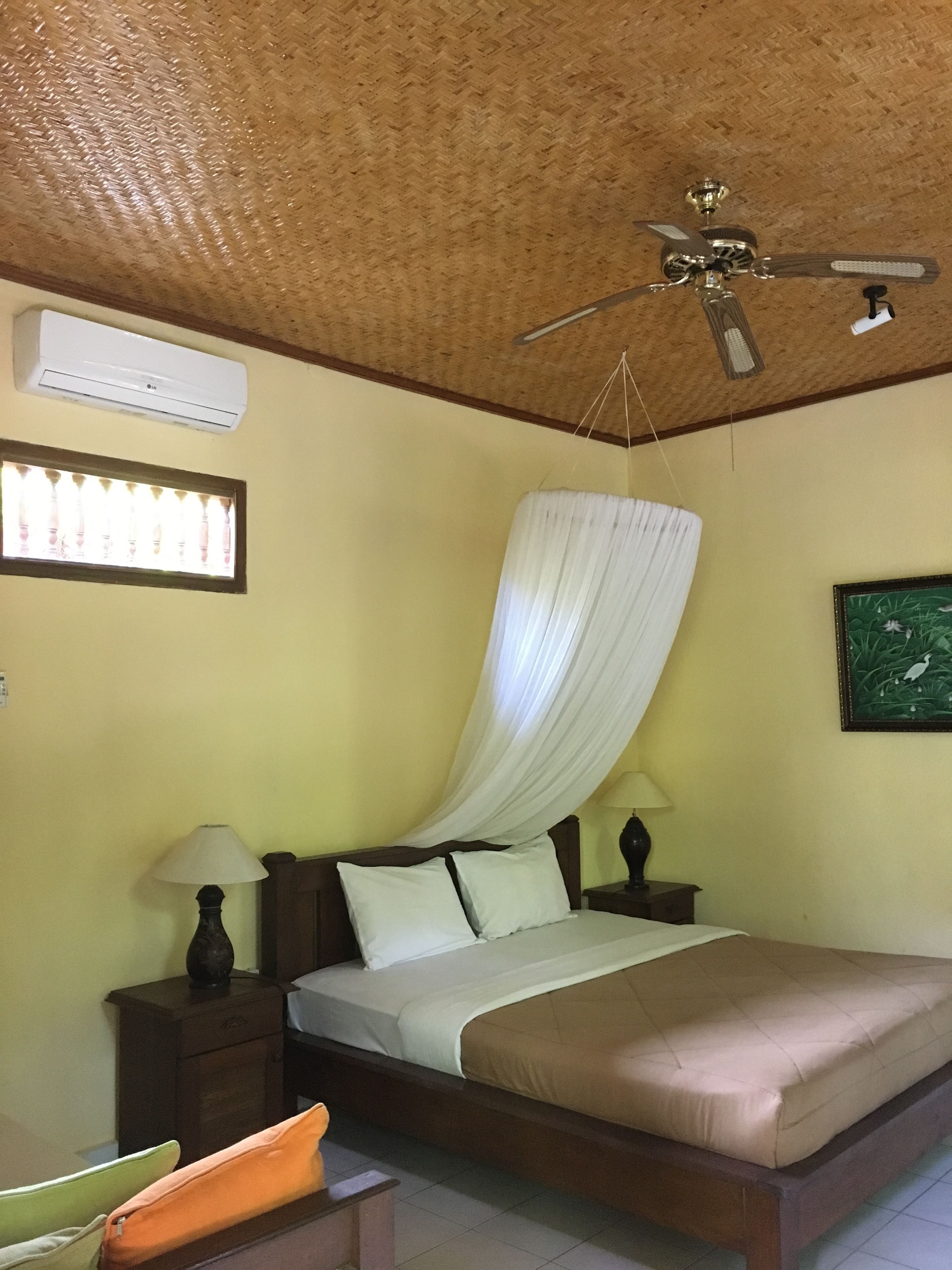 A bedroom with a bed, a ceiling fan, an air conditioning unit, a window with curtains, and a nightstand with a lamp.