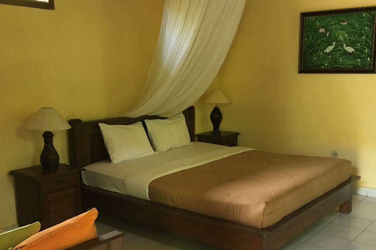 A bedroom with a bed, bedside tables, a hanging canopy, a nightstand, a lamp, a picture on the wall, and a window with blinds.