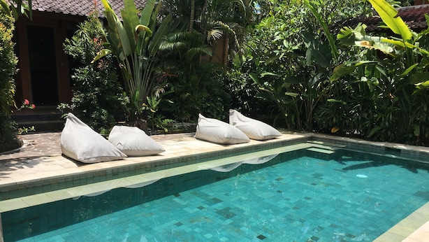 Une piscine aux eaux cristallines, bordée de carreaux, entourée d’une végétation luxuriante et agrémentée d’un patio carrelé avec des coussins blancs.